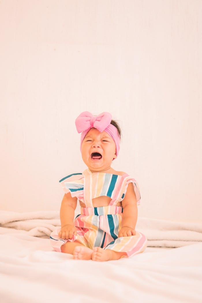A cute baby girl in a striped dress with a pink headband is crying while sitting on a bed. Soft, warm mood.